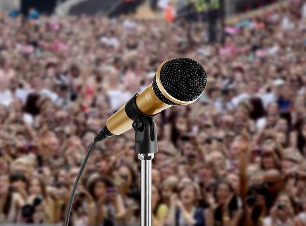 A person standing on a stage, holding a microphone and speaking to an audience.